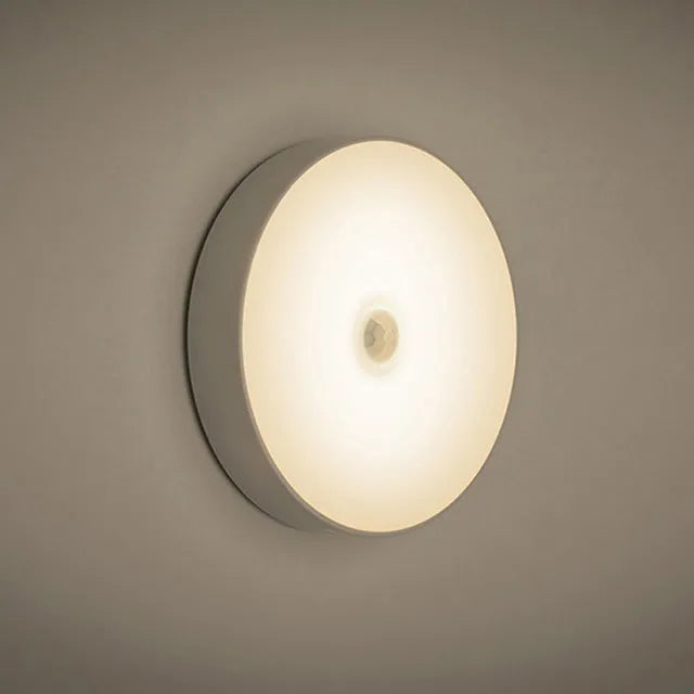 Small white circular motion sensing LED night light with adhesive backing next to a USB charging cable.