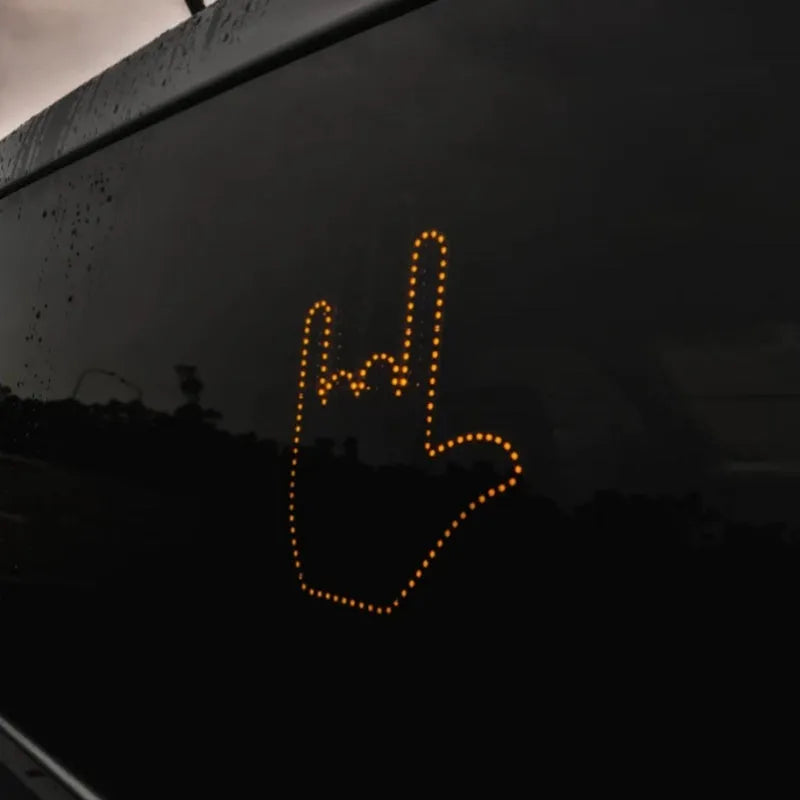 Black plastic LED gesture car light with amber LEDs showing a hand gesture, remote control, and 3M tape for mounting.