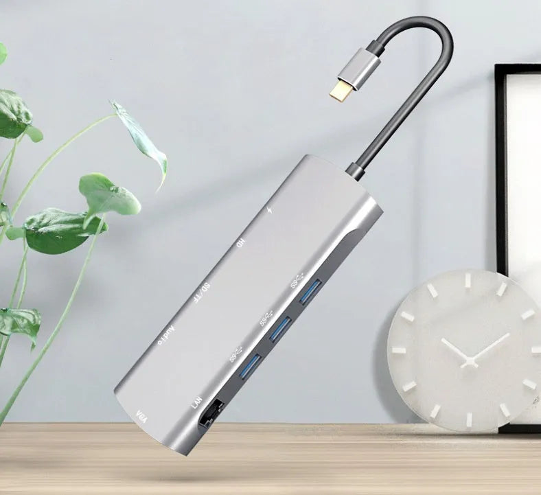 Grey aluminium USB-C docking station with HDMI, VGA, USB, and charging ports on a desk.